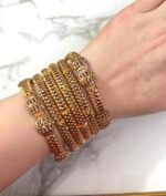 A wrist adorned with six intricately designed gold-plated mesh bangles featuring diamond-like stone clusters, set against a white marble background. The bangles showcase a braided texture and elegant beadwork, adding a luxurious and festive look.