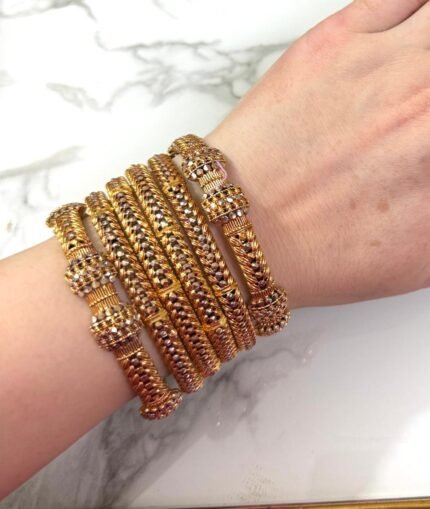 A wrist adorned with six intricately designed gold-plated mesh bangles featuring diamond-like stone clusters, set against a white marble background. The bangles showcase a braided texture and elegant beadwork, adding a luxurious and festive look.