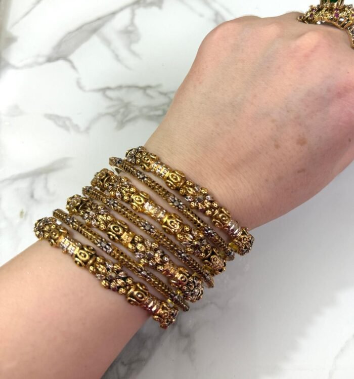 A wrist adorned with six intricately designed gold-plated bangles featuring floral motifs, beaded textures, and white stone embellishments, displayed against a white marble background.