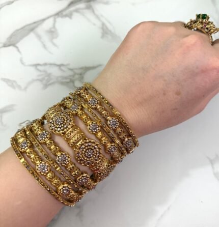 A hand wearing eight antique gold-plated bangles adorned with clustered white stones and floral patterns, displayed on a marble surface with manicured nails