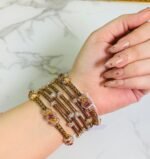 A hand adorned with a set of antique gold bangles featuring intricate dual-tone floral motifs and ruby-colored stone accents, displayed against a marble background.