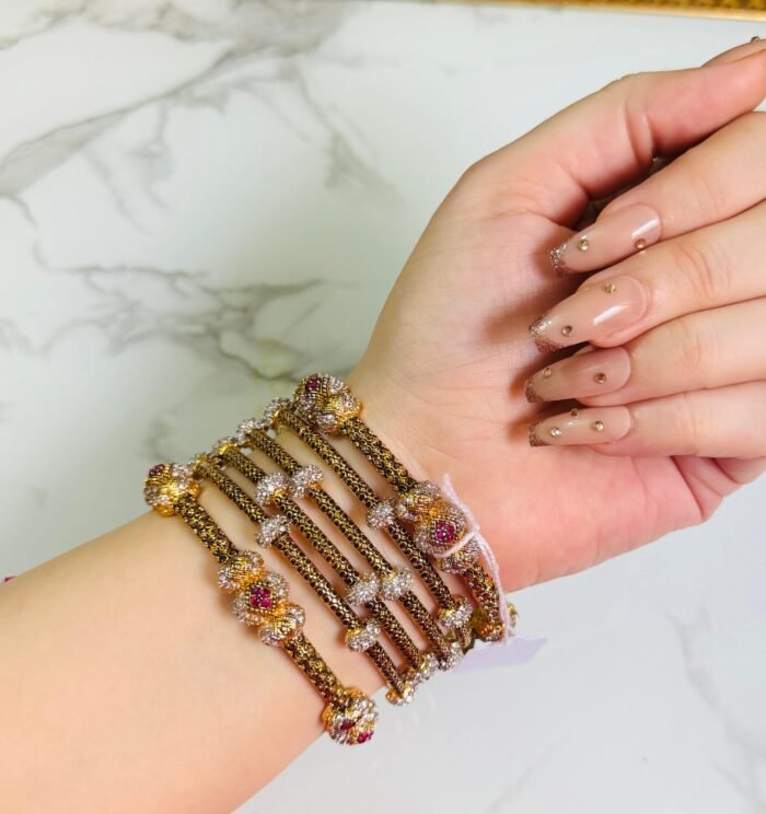 A hand adorned with a set of antique gold bangles featuring intricate dual-tone floral motifs and ruby-colored stone accents, displayed against a marble background.
