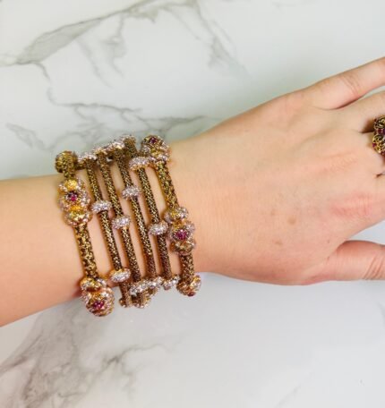 A hand adorned with a set of antique gold bangles featuring intricate dual-tone floral motifs and ruby-colored stone accents, displayed against a marble background.