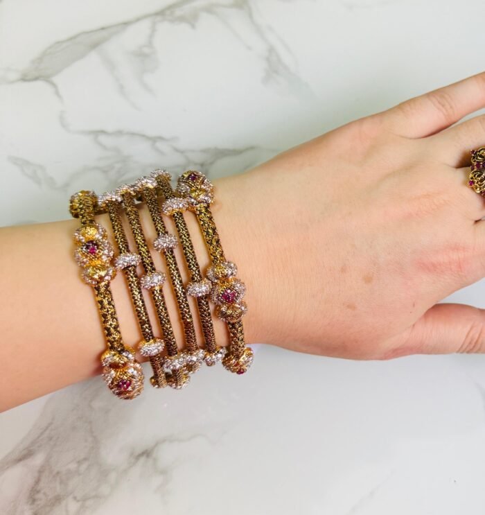 A hand adorned with a set of antique gold bangles featuring intricate dual-tone floral motifs and ruby-colored stone accents, displayed against a marble background.