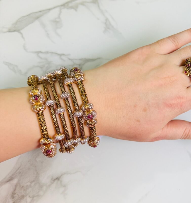 A hand adorned with a set of antique gold bangles featuring intricate dual-tone floral motifs and ruby-colored stone accents, displayed against a marble background.
