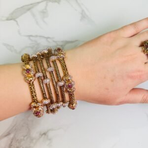 A hand adorned with a set of antique gold bangles featuring intricate dual-tone floral motifs and ruby-colored stone accents, displayed against a marble background.