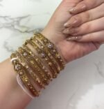 A hand adorned with a set of 8 antique gold bangles, featuring intricate floral motifs and embedded clear crystals, resting on a marble surface.