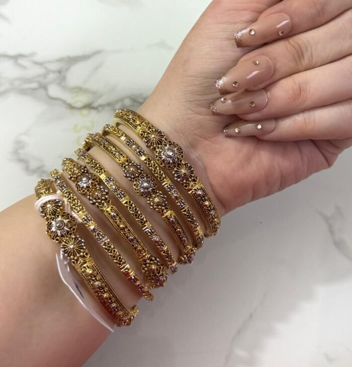 A hand adorned with a set of 8 antique gold bangles, featuring intricate floral motifs and embedded clear crystals, resting on a marble surface.