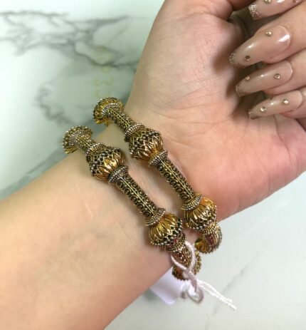 Wrist adorned with two antique-style gold bangles featuring floral motifs and detailed filigree work, set against a marble background.