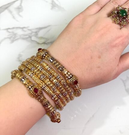 Hand wearing a traditional gold-plated bangle set with intricate detailing and red stone accents, set against a marble background.