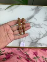 Hand holding a pair of antique-style gold bangles featuring ruby-colored center stones and intricate beaded detailing, placed over a marble surface with a pink floral fabric in the background.