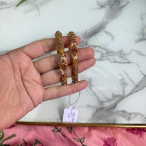 Hand holding a pair of antique-style gold bangles featuring ruby-colored center stones and intricate beaded detailing, placed over a marble surface with a pink floral fabric in the background.
