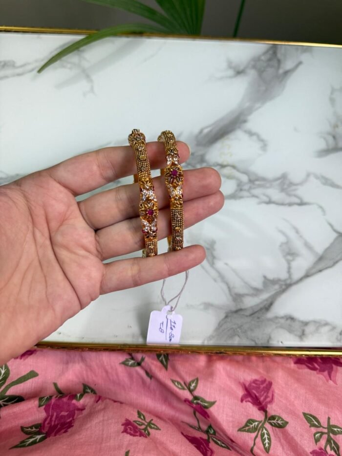 Hand holding a pair of antique-style gold bangles featuring ruby-colored center stones and intricate beaded detailing, placed over a marble surface with a pink floral fabric in the background.