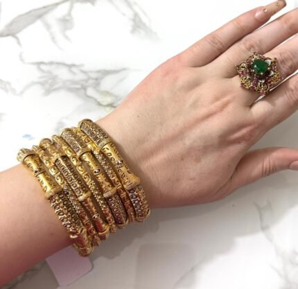 Hand wearing an elegant carved gold-plated bangle set with stone detailing and a statement green gemstone ring, placed over a marble background.