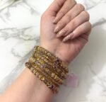 A wrist adorned with six ornate dual-tone gold bangles featuring ruby-colored stones, intricate detailing, and paired with a large green gemstone ring on a white marble background.