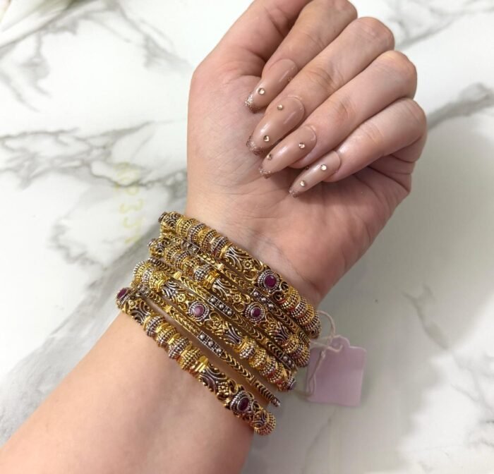 A wrist adorned with six ornate dual-tone gold bangles featuring ruby-colored stones, intricate detailing, and paired with a large green gemstone ring on a white marble background.