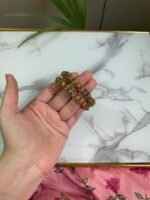 A hand holding a pair of antique gold-toned bangles with intricate floral filigree work and beaded textures, placed against a marble background with a touch of greenery.