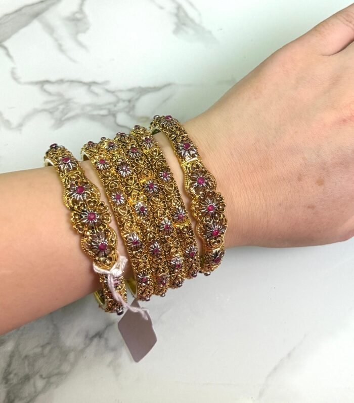 A hand wearing six antique gold bangles with intricate floral designs, featuring ruby red and white stone embellishments. The bangles are detailed with filigree work and displayed against a white marble background. A small price tag is tied to one bangle, and the overall style is traditional and ornate.