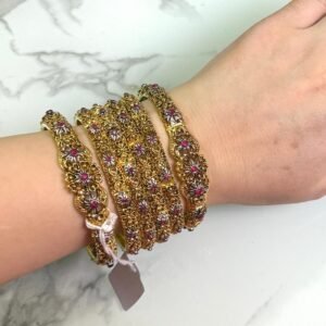 A hand wearing six antique gold bangles with intricate floral designs, featuring ruby red and white stone embellishments. The bangles are detailed with filigree work and displayed against a white marble background. A small price tag is tied to one bangle, and the overall style is traditional and ornate.