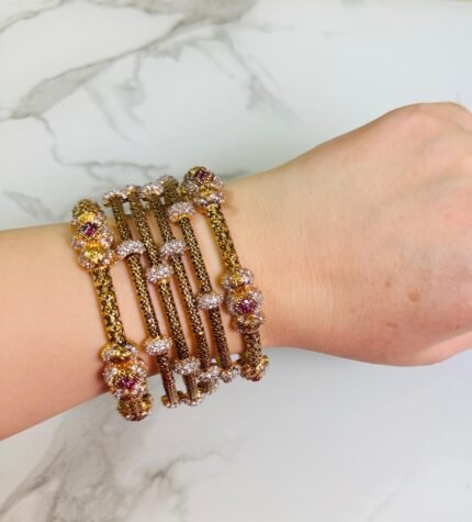 A hand adorned with a set of antique gold bangles featuring intricate dual-tone floral motifs and ruby-colored stone accents, displayed against a marble background.
