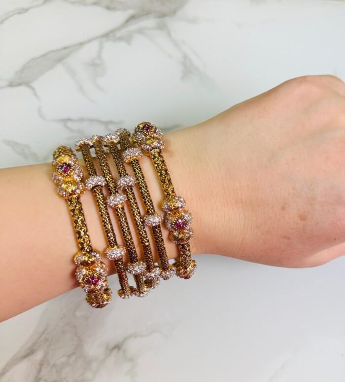 A hand adorned with a set of antique gold bangles featuring intricate dual-tone floral motifs and ruby-colored stone accents, displayed against a marble background.