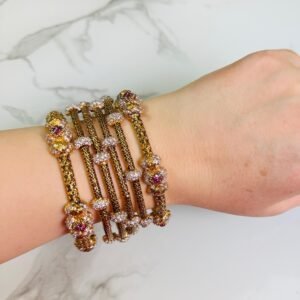 A hand adorned with a set of antique gold bangles featuring intricate dual-tone floral motifs and ruby-colored stone accents, displayed against a marble background.