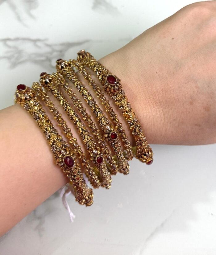 A hand adorned with a set of six antique gold bangles, each intricately designed with ornate patterns and embedded with oval-shaped deep red stones