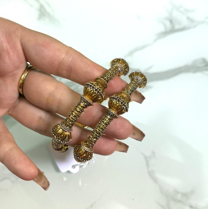 Wrist adorned with two antique-style gold bangles featuring floral motifs and detailed filigree work, set against a marble background.