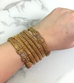 A hand wearing six gold-toned textured bangles with intricate detailing and floral designs, displayed on a white marble background