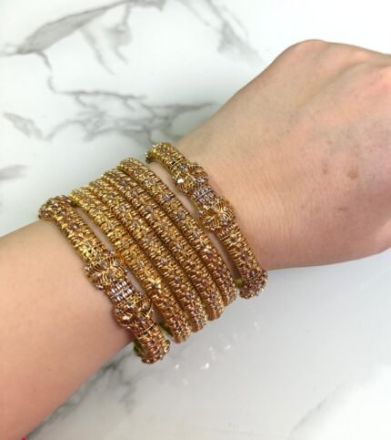 A hand wearing six gold-toned textured bangles with intricate detailing and floral designs, displayed on a white marble background