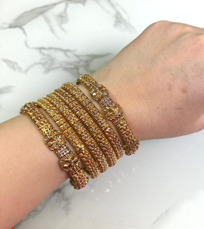 A hand wearing six gold-toned textured bangles with intricate detailing and floral designs, displayed on a white marble background