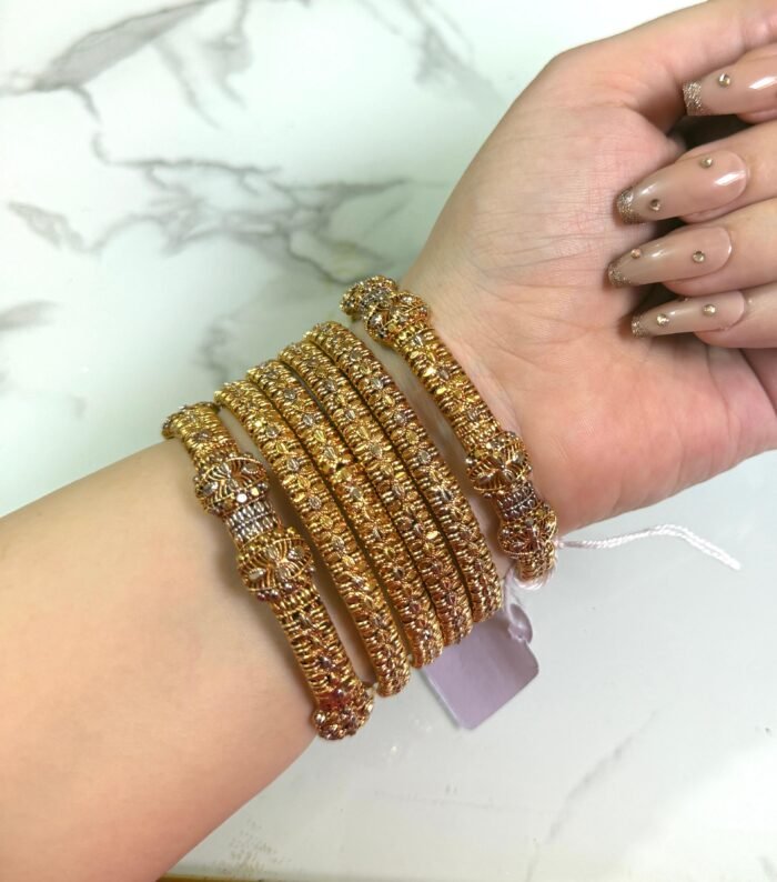 A hand wearing six gold-toned textured bangles with intricate detailing and floral designs, displayed on a white marble background