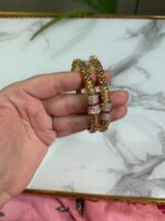A hand holding two gold-toned bangles with intricate floral patterns and sparkling stone-studded accents in silver and ruby hues, displayed against a marble background.