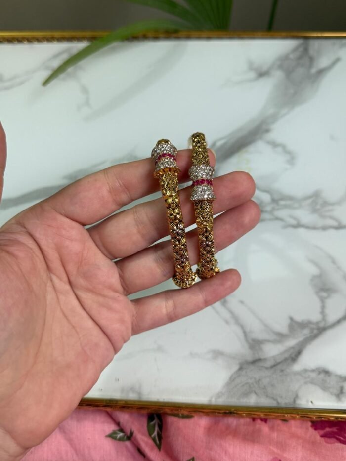 A hand holding two gold-toned bangles with intricate floral patterns and sparkling stone-studded accents in silver and ruby hues, displayed against a marble background.