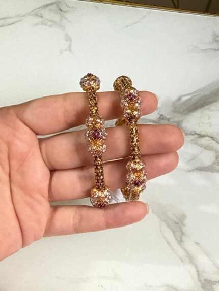 A pair of ornate gold bangles adorned with clusters of multicolor stones in shades of red, pink, and white, held in a hand over a marble background.