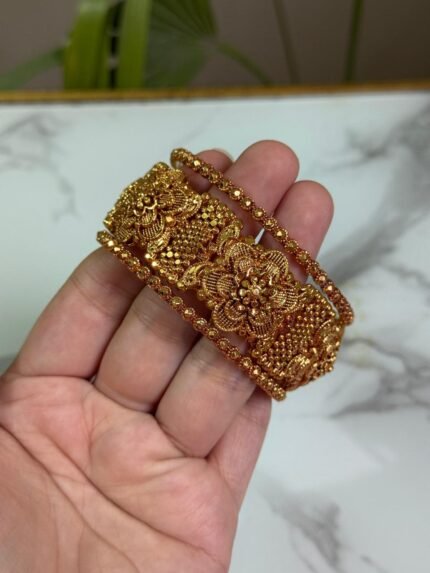 A wide gold kada bangle with intricate floral engravings and beadwork, held in a hand against a marble background.