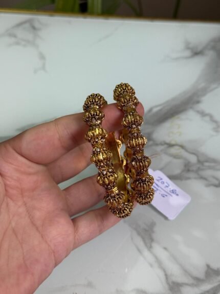 A pair of antique gold plated kada bangles with intricate bulb-shaped temple motifs held in a hand over a marble surface.