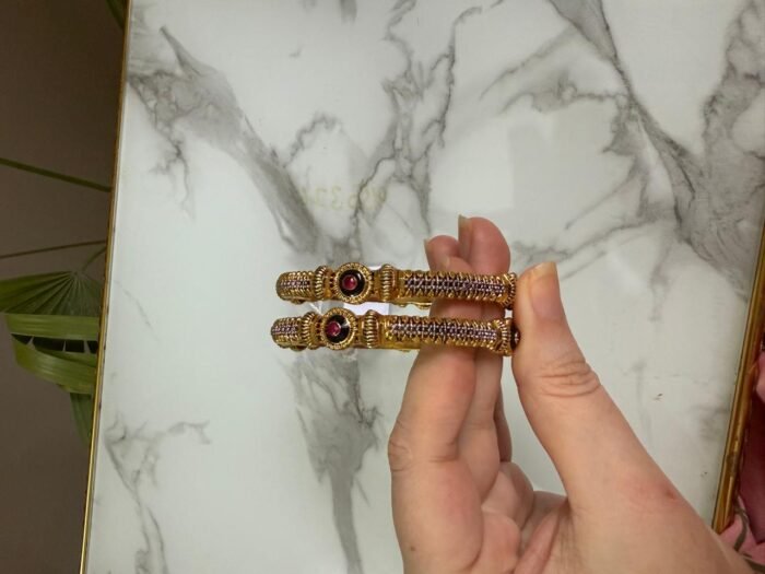 A pair of antique gold bangles with intricate textured design and round pink gemstone centers, held in hand against a marble backdrop.