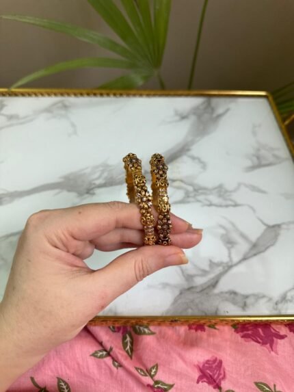 A pair of antique-style gold bangles featuring intricate floral and filigree patterns, held in hand against a marble background.