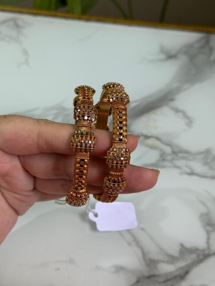 A pair of traditional gold bangles with rope-texture design and ornate bead detailing, held in hand over a marble surface.