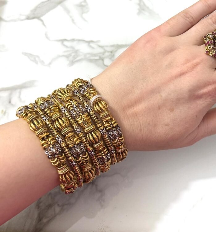Hand wearing a grand antique gold-plated bangle set with intricate traditional designs and silver detailing, displayed on a marble background.
