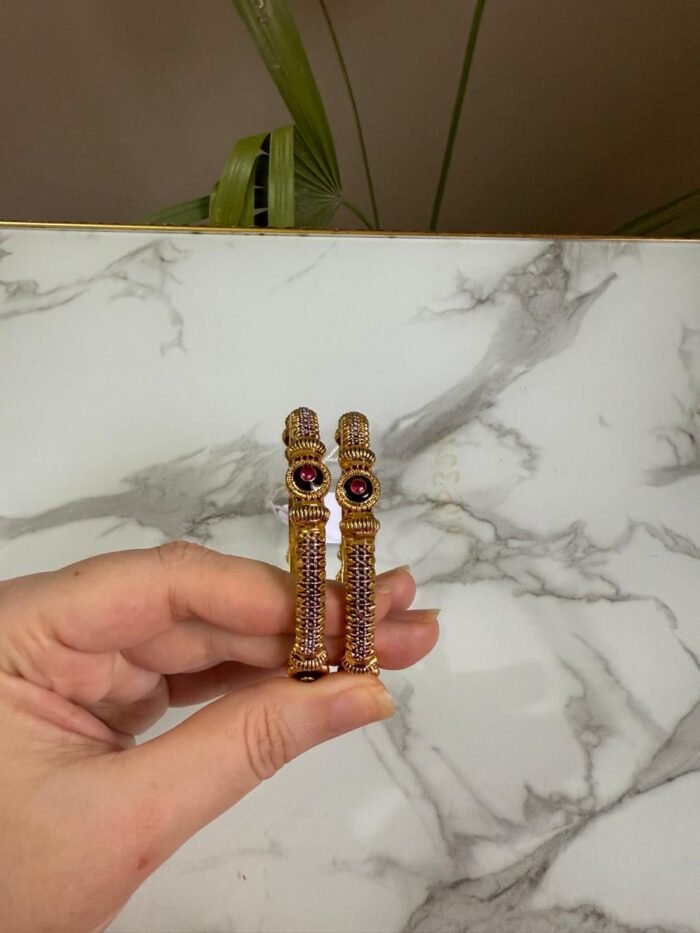 A pair of antique gold bangles with intricate textured design and round pink gemstone centers, held in hand against a marble backdrop.