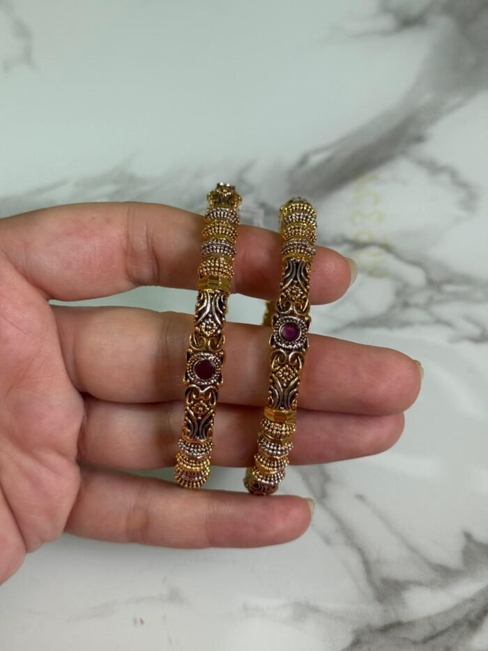 A close-up of a hand holding a pair of gold-plated ethnic bangles with intricate filigree patterns and ruby red stone embellishments, against a marble background.
