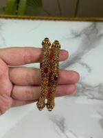 A hand holding a pair of antique-finish gold bangles with intricate floral carvings and red oval center stones, displayed against a marble background.