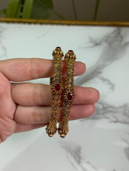 A hand holding a pair of antique-finish gold bangles with intricate floral carvings and red oval center stones, displayed against a marble background.