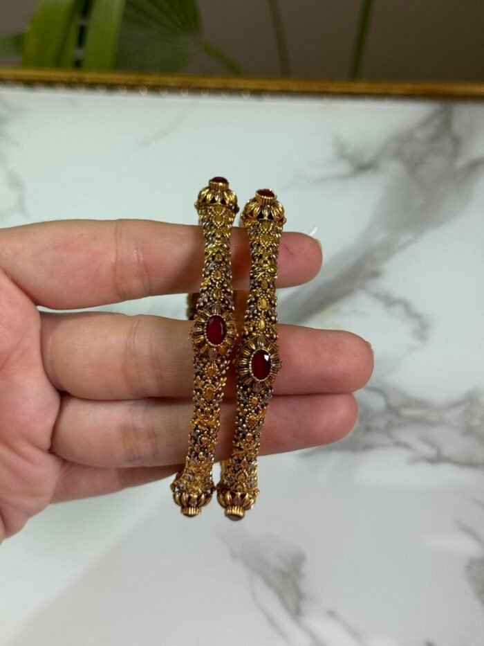 A hand holding a pair of antique-finish gold bangles with intricate floral carvings and red oval center stones, displayed against a marble background.
