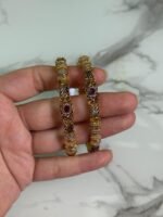 A close-up of a hand holding a pair of gold-plated ethnic bangles with intricate filigree patterns and ruby red stone embellishments, against a marble background.