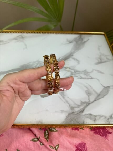 A pair of antique-style gold bangles featuring intricate floral and filigree patterns, held in hand against a marble background.