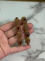 A pair of ornate antique gold bangles with large red oval gemstones and leaf-patterned goldwork, held in a hand over a marble background.