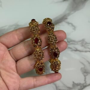A pair of ornate antique gold bangles with large red oval gemstones and leaf-patterned goldwork, held in a hand over a marble background.
